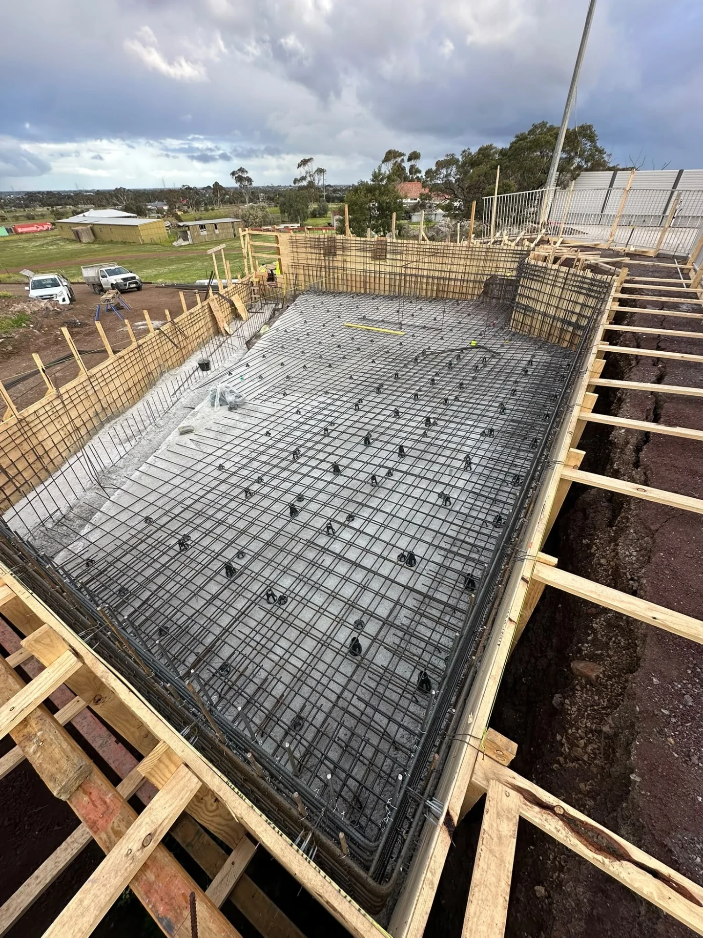 Large pool floor with layered reinforcement on bar chairs, surrounded by tall formwork walls and timber bearers over an open trench.