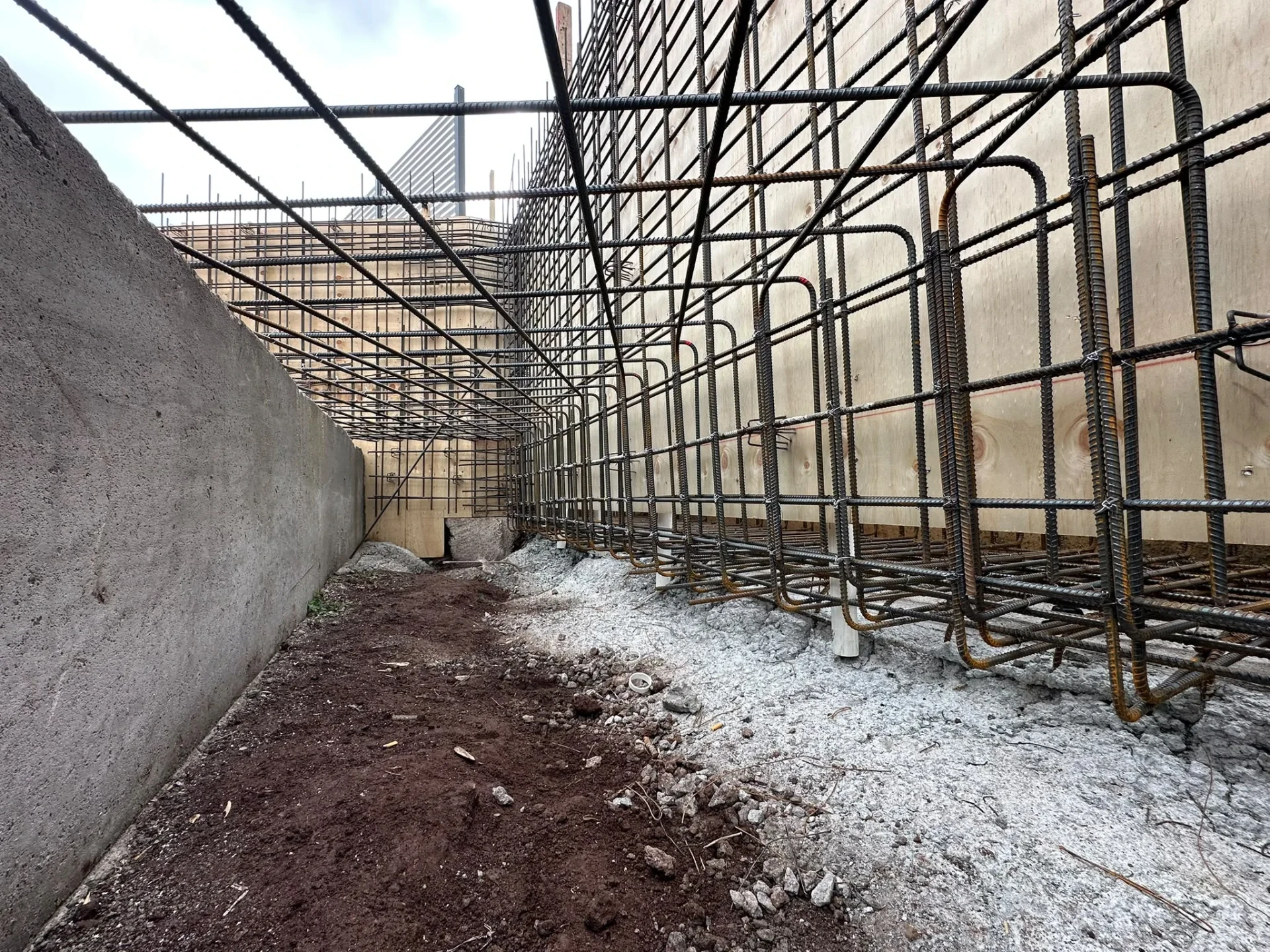 Perimeter trench between a concrete wall and pool formwork, with looped reinforcement cages and service pipe stub-ups.