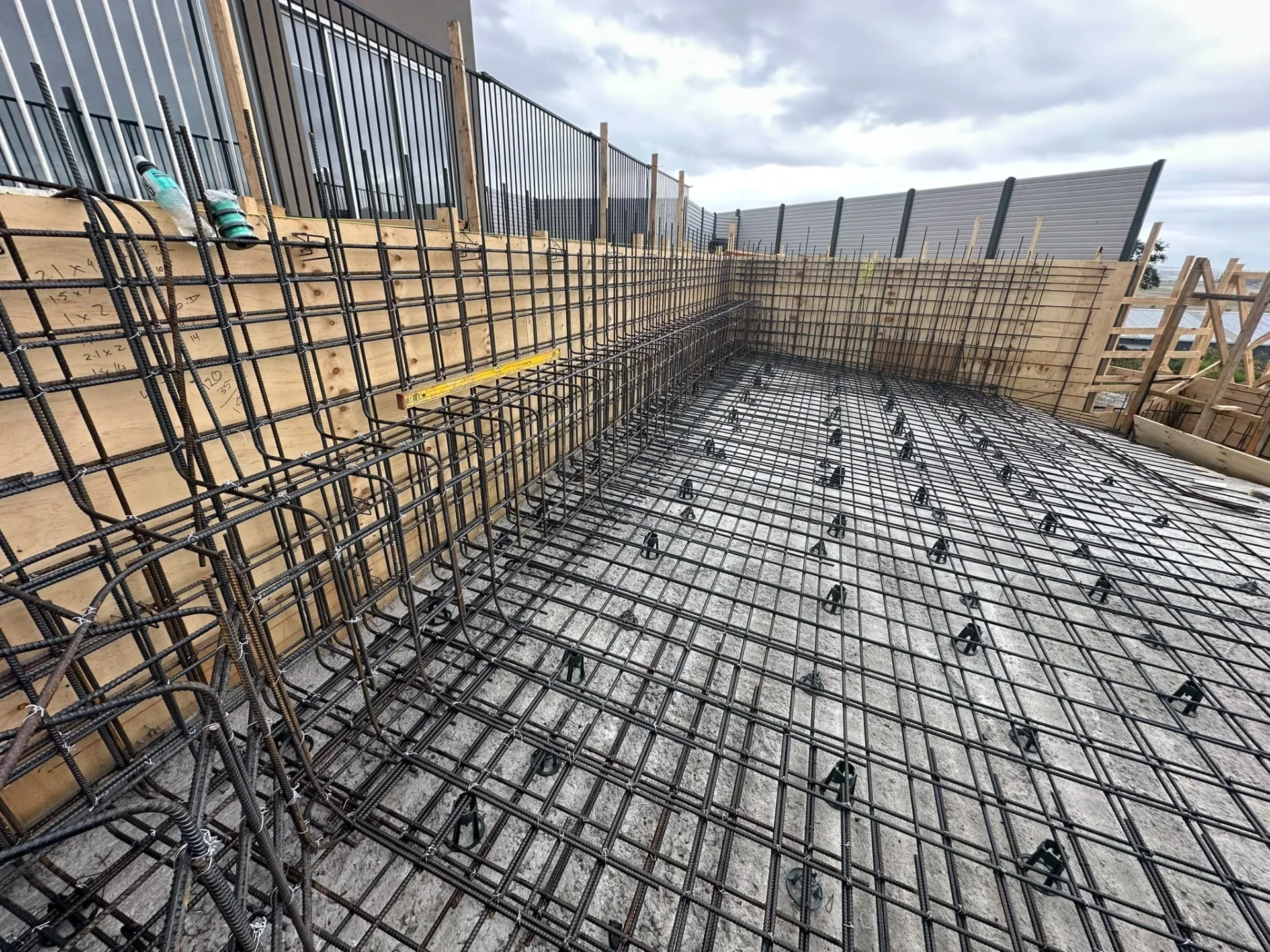 Pool floor reinforcement and wall steel inside plywood formwork alongside an existing building and boundary fencing.