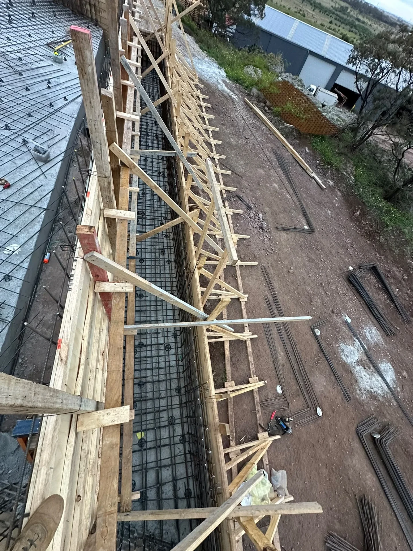 Timber formwork wall with raking braces along one side of a pool, with bent reinforcement bar laid out on the ground.