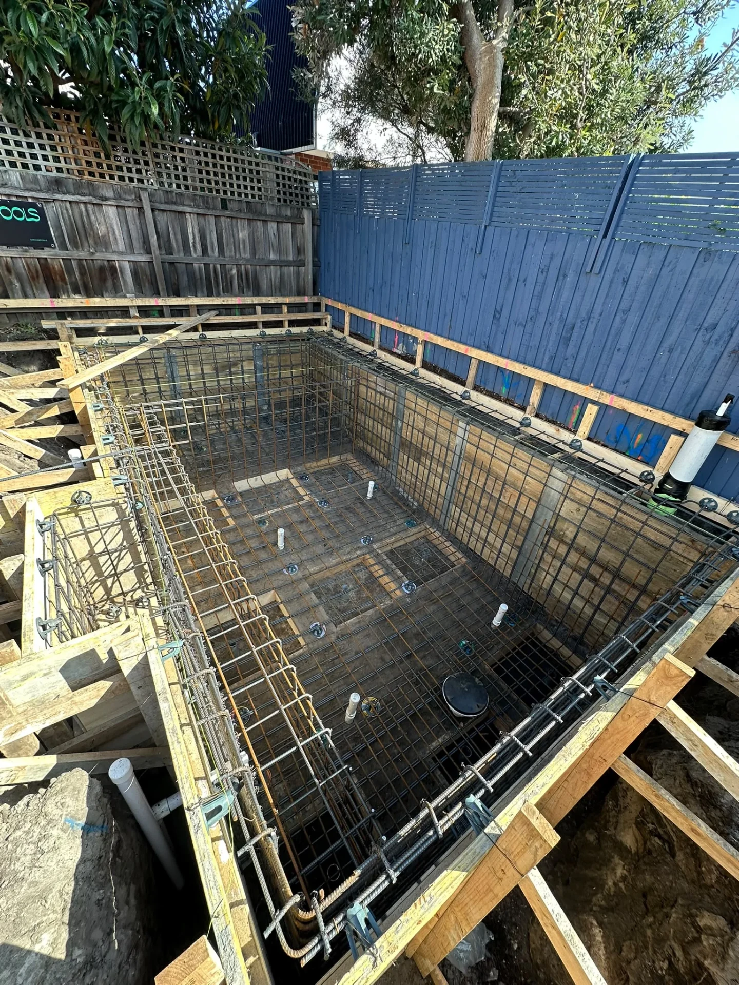 Rectangular pool cavity in a fenced backyard with wall reinforcement cages, timber formwork and plumbing stub-ups.