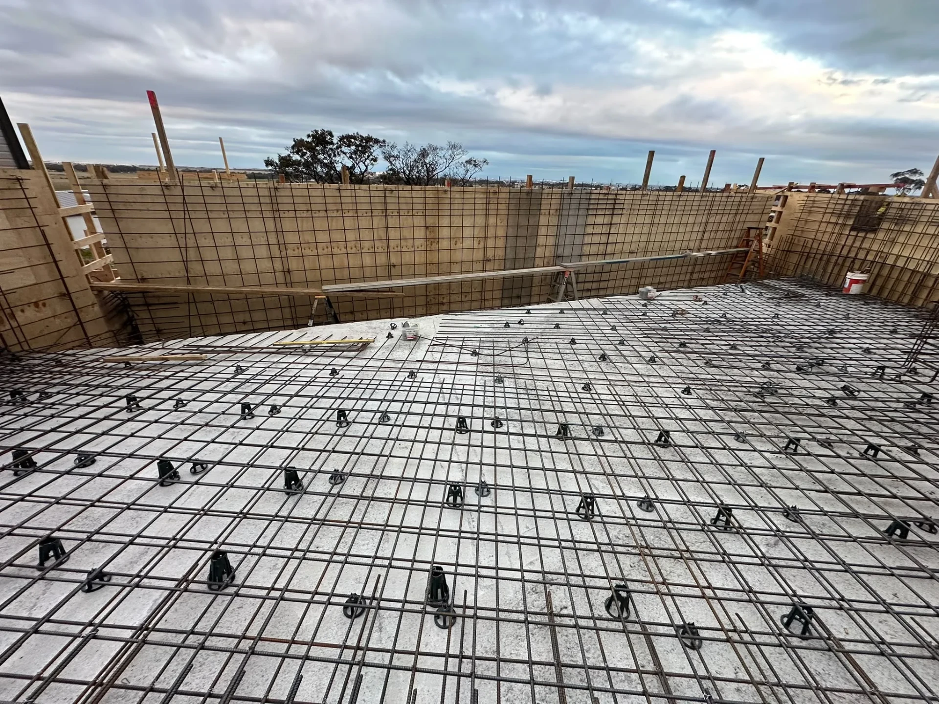 Pool floor covered with tied steel mesh on bar chairs inside plywood formwork walls, under an overcast sky.