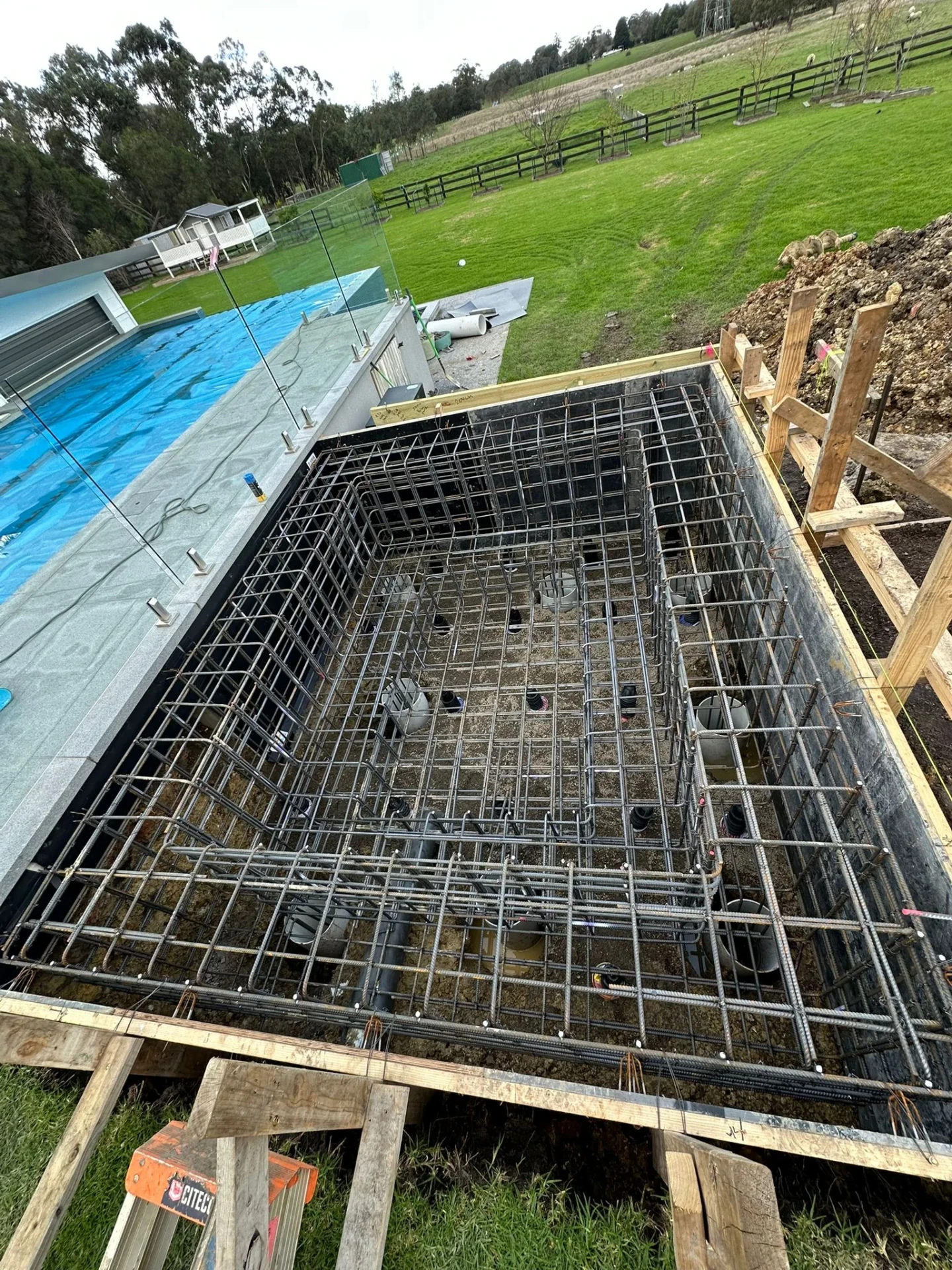Rectangular reinforcement cage set in an excavation beside a covered pool with glass fencing, on a rural property.