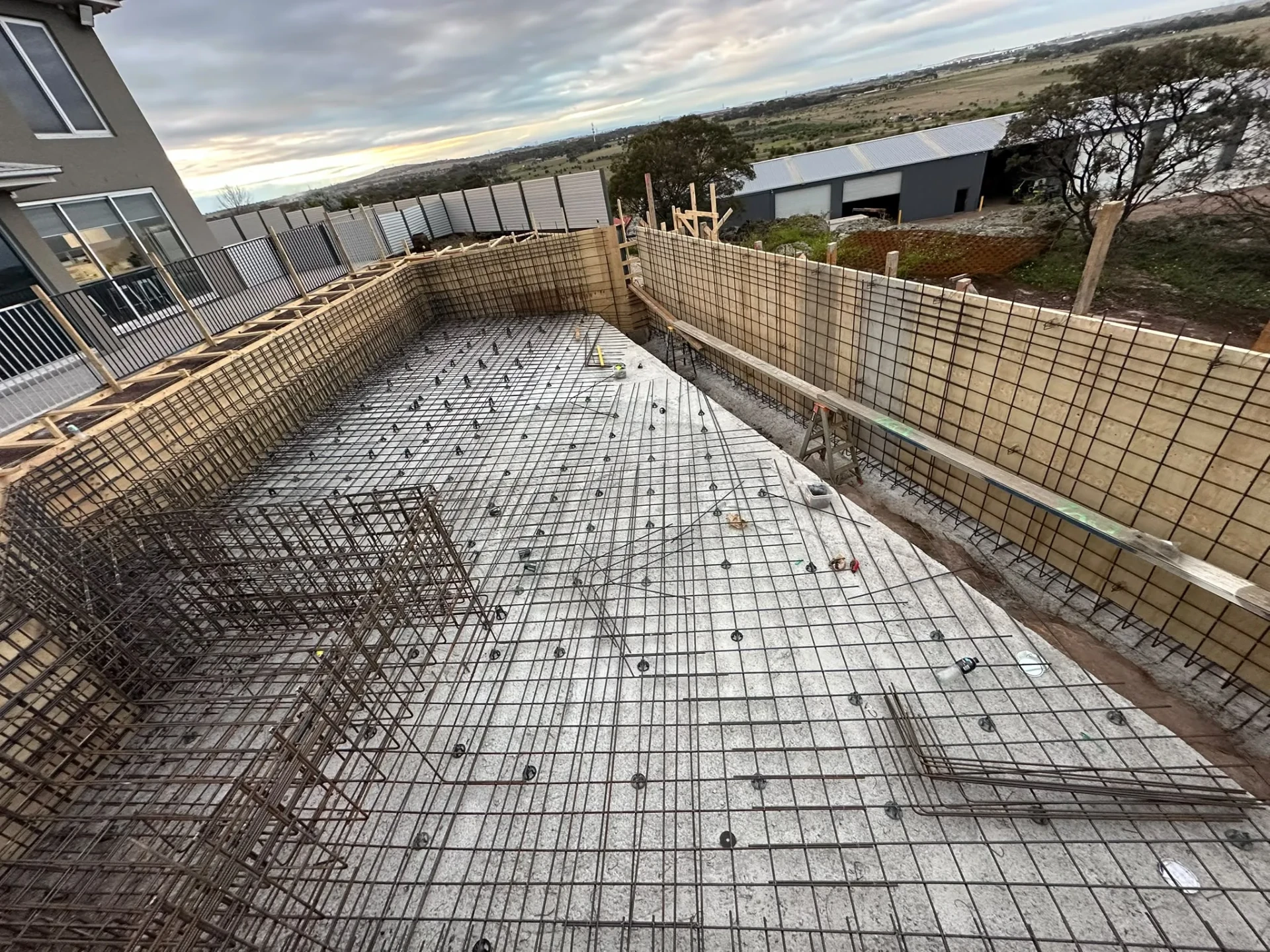 Long pool cavity beside a two-storey house with braced timber formwork walls, wall mesh and floor reinforcement.