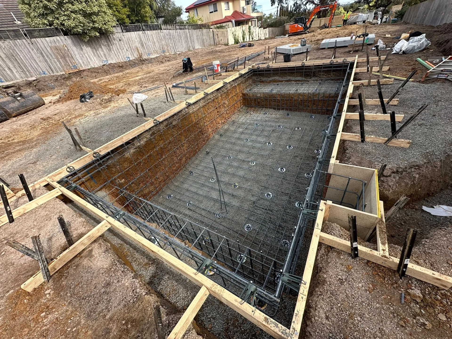 Rectangular pool excavation with floor mesh on bar chairs, perimeter formwork with steel props and an excavator on site.