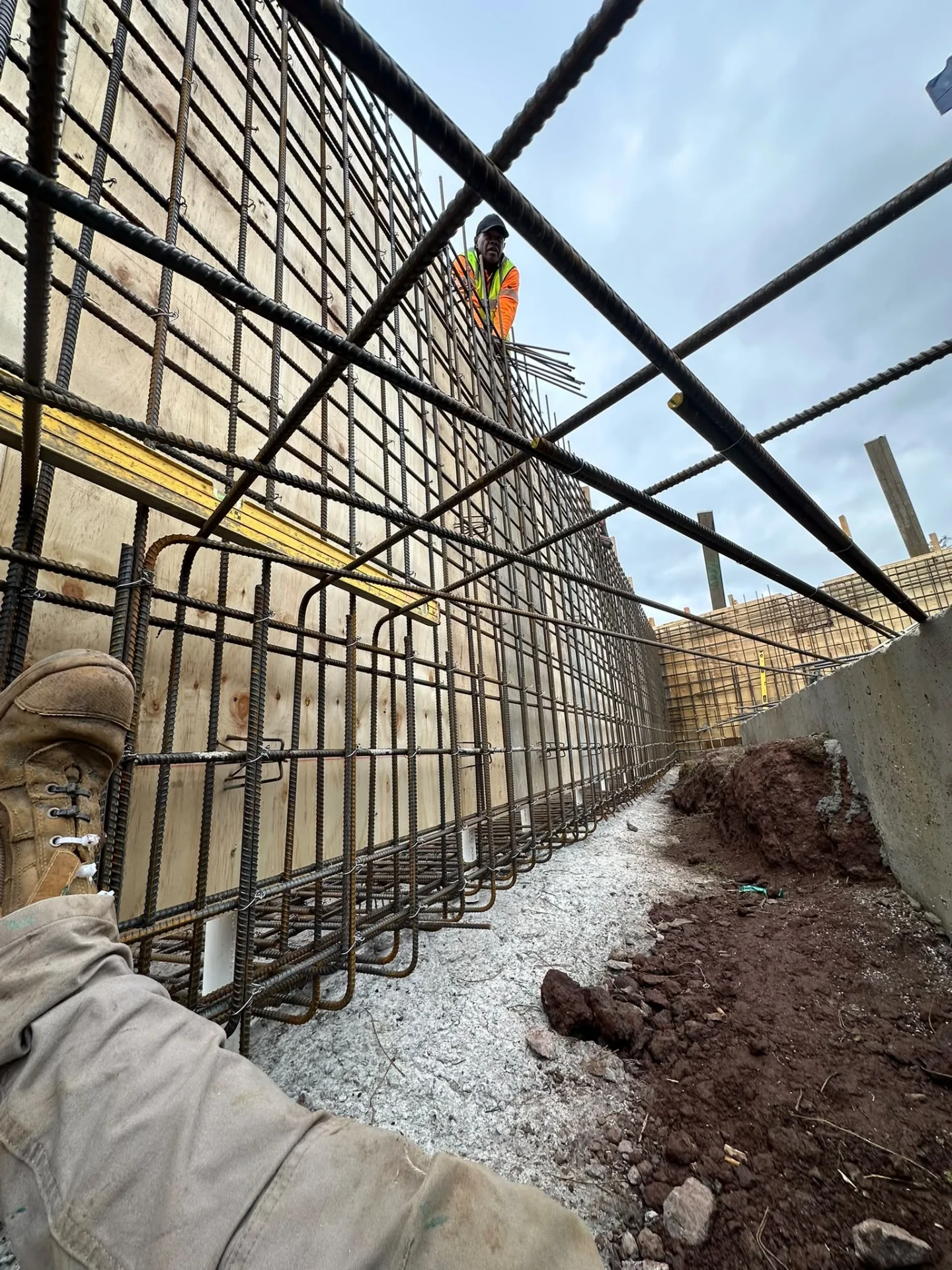 Low view along a formwork wall with vertical and horizontal reinforcement bars tied in place and a worker above the wall.
