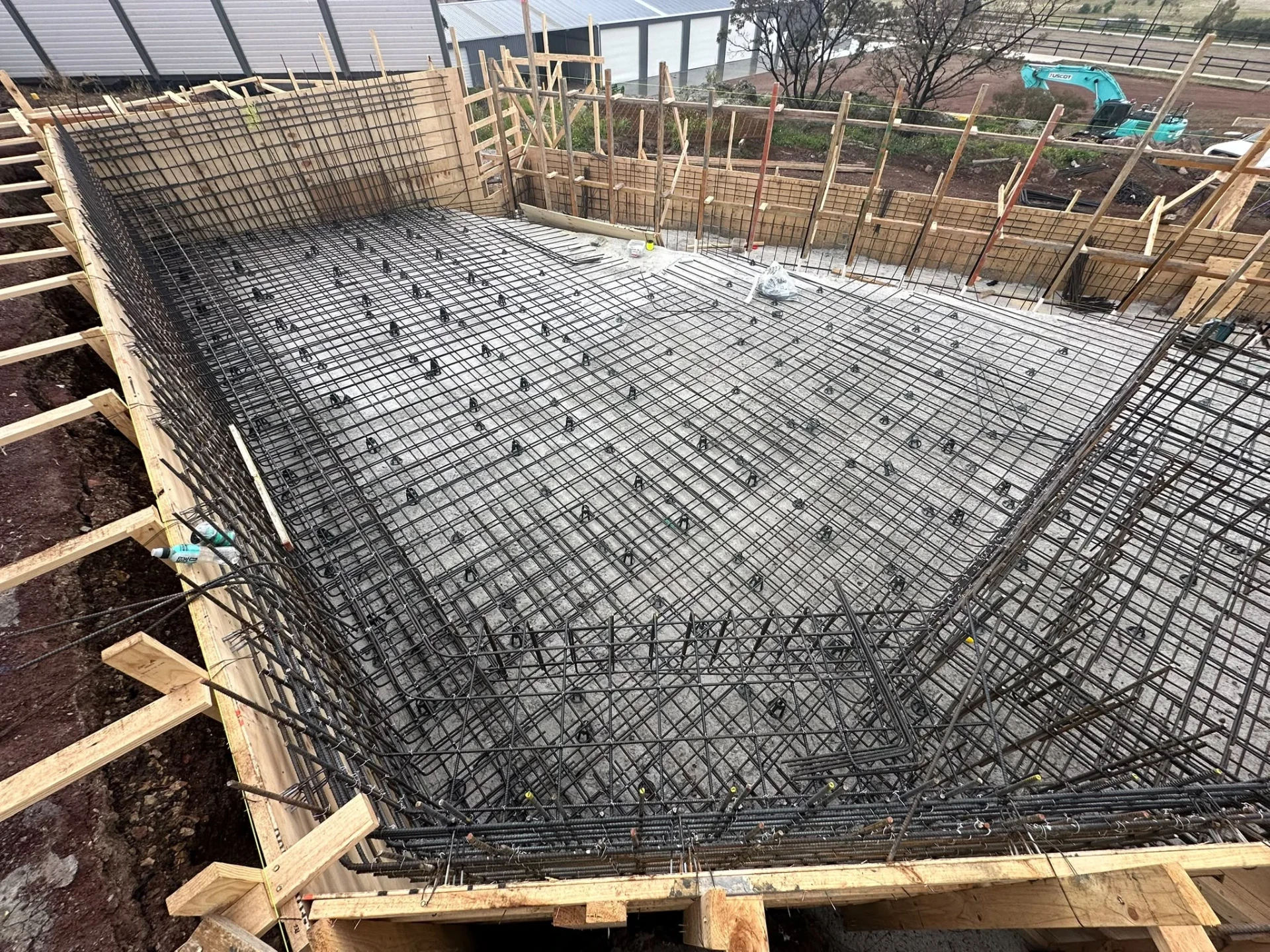Elevated view of a rectangular pool with floor reinforcement, formed and braced perimeter walls and an excavator on site.