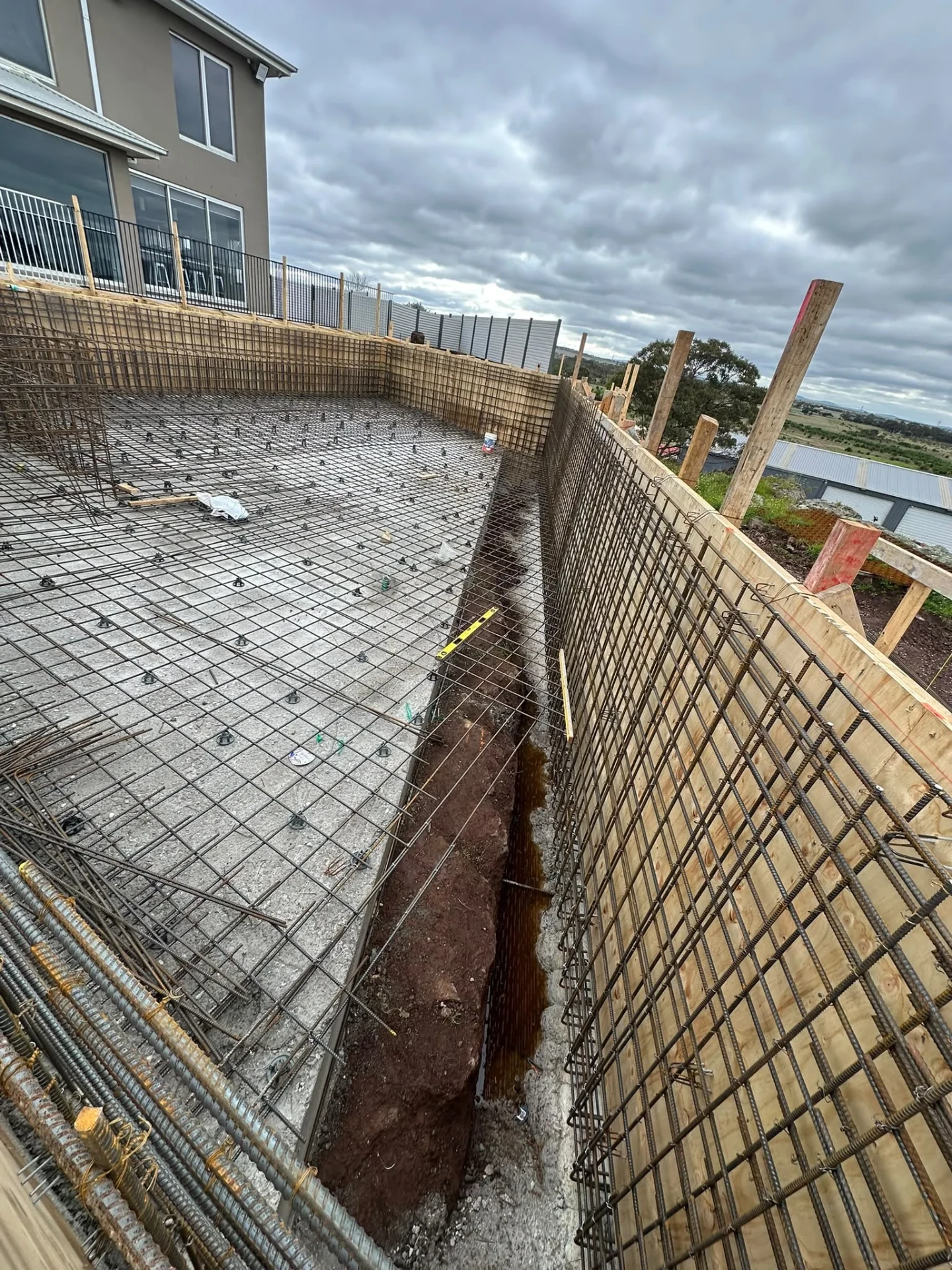 Pool floor reinforcement with an open perimeter trench and mesh wall panels, next to a two-storey house.